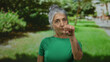 © Krakenimages.com - Grey-haired senior woman in green gestures listening and knocking in outdoor park setting, surrounded by greenery and sunlight, conveying curiosity and communication.