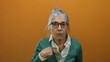 © Krakenimages.com - Woman with grey hair and glasses pointing at herself, wearing a green cardigan against an orange background, expressing surprise and curiosity.