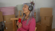 © Krakenimages.com - Woman with grey hair enjoying music in new home surrounded by packed boxes and holding red smartphone, wearing headphones and pink sweater in living room.