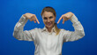 © Krakenimages.com - Young blonde woman in a white shirt gestures against a vivid blue background, exuding confidence and charm while pointing with both hands.