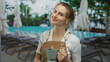 © Krakenimages.com - Woman in apron holding coffee maker by resort pool with palm trees and lounge chairs in background on sunny day