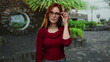 © Krakenimages.com - Woman adjusting glasses with green plants behind on a city street, showcasing her red hair and casual red sweater.