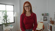 © Krakenimages.com - Woman with red hair smiling in a cozy bedroom, wearing glasses and a red sweater, standing indoors with a lamp and baby items in the background, exuding warmth and charm.
