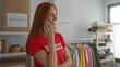 © Krakenimages.com - Woman talking on phone in charity donations room, wearing red volunteer shirt, redhead smiling with clothing and supplies on shelves.