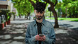 © Krakenimages.com - Young man with phone in urban street setting, looking slightly displeased, wearing denim jacket with trees and buildings in the background on a sunny day.