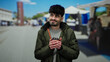 © Krakenimages.com - Young man smiling with coffee cup in hands standing at outdoor market street under a clear blue sky, wearing a green jacket, capturing a relaxed and happy moment in a vibrant urban setting.