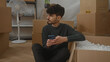 © Krakenimages.com - Young man sitting in new home surrounded by cardboard boxes using a smartphone in the living room, depicting a moment of relaxation amidst a move, with a thoughtful expression.