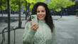 © Krakenimages.com - Woman smiling outdoors taking selfie with smartphone in urban street setting featuring green trees and sweater