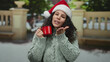 © Krakenimages.com - Hispanic woman in santa hat blows kiss holding red mug on festive outdoor city street background conveying holiday cheer and warmth.
