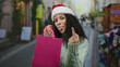 © Krakenimages.com - Young woman wearing santa hat and sweater makes rude gesture with middle finger while holding shopping bag on lively city street during holiday season