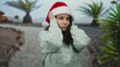 © Krakenimages.com - Woman outdoors wearing santa hat in desert, holding head in frustration, surrounded by palm trees, expressing stress during holiday season.