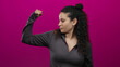 © Krakenimages.com - Young hispanic woman flexes her arm confidently against an isolated pink background wall, emphasizing strength, empowerment, and determination.