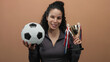 © Krakenimages.com - Woman smiling holding soccer ball and trophy against brown wall background symbolizing victory and success in sports achievement.