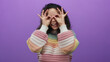 © Krakenimages.com - Woman smiling wearing colorful sweater playfully posing with her hands forming glasses against an isolated purple background expressing joy and creativity.