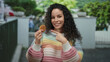 © Krakenimages.com - Woman smiling outdoors wearing colorful sweater holding cookie on street with trees in background, showcasing cheerful and relaxed mood in open urban environment.