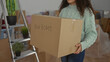 © Krakenimages.com - Hispanic woman holding box in new home interior living room symbolizing moving and beginning, surrounded by plants and unpacked furniture, wearing warm sweater with happy expression.