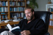 © Milos - A man with a well-groomed beard smiles warmly while reading a book in a contemporary, comfortable chair, showcasing a harmonious blend of relaxation and intellectual engagement.