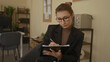© Krakenimages.com - Young woman in glasses writing in a notebook at a modern office desk, surrounded by shelves and plants, conveying focus and professionalism in an interior workplace setting.