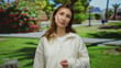 © Krakenimages.com - Young woman standing in a sunny outdoor park with blooming flowers and lush greenery, wearing a casual white sweater, enjoying a peaceful afternoon in nature.