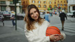 © Krakenimages.com - Young woman holding basketball in urban street setting with vibrant city background and people walking around, highlighting outdoor activity and youthful energy