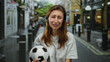 © Krakenimages.com - Woman in a city street holding a soccer ball with a sad expression, depicting emotion, outdoor scene, wearing casual white clothing, surrounded by urban cityscape.