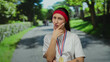© Krakenimages.com - Young woman with medal thinking in a sunlit park, wearing a red headband and white shirt, surrounded by greenery and path, outdoor pose inspiring fitness and achievement goals.