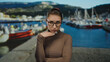 © Krakenimages.com - Woman pondering outdoor near a port with boats in the background, wearing glasses and brown top, with mountains visible, suggesting a thoughtful moment.