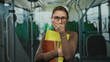 © Krakenimages.com - Young woman standing inside a bus with folders and books, covering her mouth while wearing glasses and a brown outfit, suggesting a sense of surprise or contemplation.