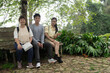 © Freddy Chandra - Portrait of a happy Asian family of three sitting on a wooden bench in a lush tropical garden at a nature park
