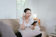 © Kieng - A young Asian woman relaxes on a sofa at home, wearing wireless headphones while reading a book, enjoying a calm moment of learning, entertainment, and modern digital lifestyle.