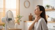 © ALLmost - A young Asian woman feeling the heat and fanning herself with a portable electric fan in a sunlit room.