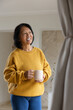 © wavebreak3 - Senior Asian woman holding large ceramic mug smiling gazing right in living area wearing sweater