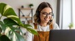 © A R - Woman wearing headset and working on laptop in a bright office environment with plants nearby