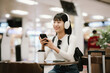 © Witoon - A young Asian woman wearing headphones sits at an airport café, using her smartphone beside a suitcase and coffee cup, reflecting modern travel, digital lifestyle, and waiting time.