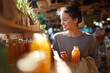 © EricMiguel - Smiling young woman selecting fresh bottled juices at a cozy organic market — holding glass bottles and a reusable bag in warm sunlight