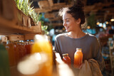 Smiling young woman selecting fresh bottled juices at a cozy organic market — holding glass bottles and a reusable bag in warm sunlight