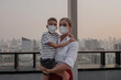 © blackday - Mother holding young child while both wear protective face masks on rooftop with city skyline