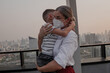 © blackday - Mother holding young child while both wear protective face masks on rooftop with city skyline