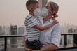 © blackday - Mother holding young child while both wear protective face masks on rooftop with city skyline