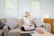 © NANKHAMMAI - A young woman is relaxing on the sofa at home, smiling, holding a cup of tea, enjoying a peaceful weekend. The warm and cozy atmosphere of the living room, a relaxed lifestyle, and a blissful daydream