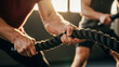 © vladnikon - A dynamic close-up of hands gripping battle ropes during an intensive gym workout, highlighting muscular strain and functional fitness with a blurred training partner in the background