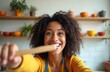 © Viktor - Smiling woman holds wooden spoon in kitchen. She films herself cooking meal, preparing healthy food. Young adult enjoys domestic life at home.
