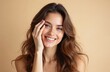 © Viktor - Smiling woman touches her face with hand. She has long wavy brown hair and perfect white teeth. Her eyes are brown and she looks happy and confident. Image is on a plain neutral background.