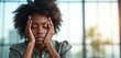 © Viktor - Black woman holds head with hands eyes closed. Stressed professional feels headache from work overload. Female employee experiences burnout and anxiety at desk. Mental health issue in office.