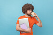 © khosrork - Portrait of tired man office worker with Afro hairstyle in orange T-shirt standing with laptop and paper notebook, drinking coffee needs energy for work. Indoor studio shot isolated on blue background