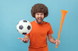 © khosrork - Portrait of excited crazy man with Afro hairstyle wearing orange T-shirt holding soccer ball and horn, screaming, cheering, watching football match. Indoor studio shot isolated on blue background.
