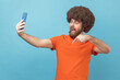 © khosrork - Portrait of excited man with Afro hairstyle posing at phone camera, making selfie or having video call, pointing down asking to subscribe. Indoor studio shot isolated on blue background.