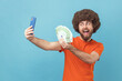 © khosrork - Portrait of cheerful man with Afro hairstyle wearing orange T-shirt having video call or livestream, boasting euro banknotes to subscribers. Indoor studio shot isolated on blue background.