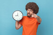 © khosrork - Portrait of man with Afro hairstyle wearing orange T-shirt holding wall clock in hands and pointing to camera at you, time management, you are late. Indoor studio shot isolated on blue background.