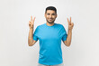© khosrork - Man wearing blue T- shirt standing shows peace v sign, smiles toothily, enjoys nice day gestures victory symbol, has good mood carefree expression. Indoor studio shot isolated on gray background.